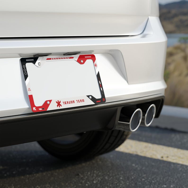 May include: A rectangular license plate frame featuring a white, red, and black geometric design with the text "TRAUMA TEAM" printed at the bottom. The frame includes small circular icons and linear patterns along the top and side borders.
