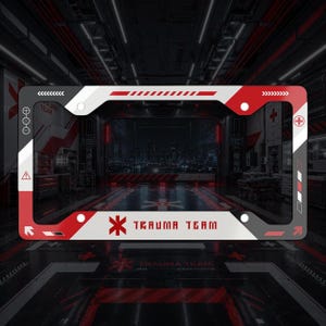 May include: A rectangular license plate frame featuring a white, red, and dark gray color scheme with geometric patterns. The frame displays the text "TRAUMA TEAM" in a bold, stylized font, accompanied by a red medical cross symbol and various icons.
