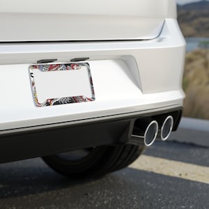 K&ouml;nnte beinhalten: Ein Nummernschildrahmen mit einem farbenfrohen, komplizierten Design mit roten, schwarzen und wei&szlig;en Mustern. Der Rahmen ist an der R&uuml;ckseite eines wei&szlig;en Autos montiert, wobei die Auspuffrohre darunter sichtbar sind.