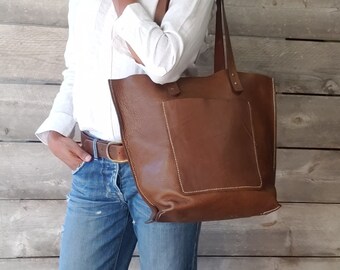 Puede incluir: Un bolso tote de cuero marrón con un bolsillo frontal grande y un cierre de cremallera superior. El bolso lo lleva una persona que lleva una camisa blanca y pantalones vaqueros azules.