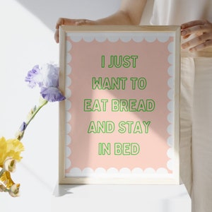 May include: A framed print with a pink background and white scalloped border. The text reads "I just want to eat bread and stay in bed" in green.