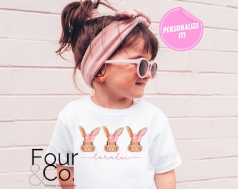 May include: White t-shirt featuring three brown bunnies with pink flowers on their heads. The bunnies are arranged in a row with the text "loralei" underneath. The shirt is being worn by a child with brown hair and a pink headband.