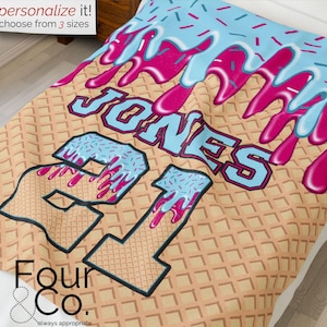 May include: A personalised blanket with a waffle cone pattern background. The blanket features the name "JONES" and the number "21" in a blue and pink ice cream design. The top of the blanket has a blue ice cream drip design with sprinkles. The text "personalize it! choose from 3 sizes" is in the top left corner.