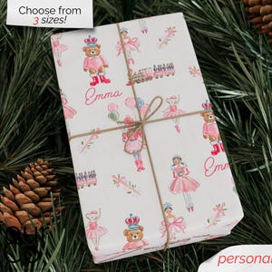 May include: A wrapped gift with a white background, featuring a repeating pattern of pink ballerinas, teddy bears, and toy trains. The name "Emma" is written in pink script. The gift is tied with twine and surrounded by pine needles and pine cones.