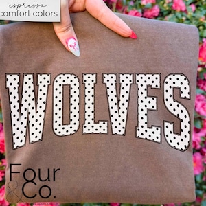 May include: A brown t-shirt with the word "WOLVES" in large, white, polka-dotted letters. The text "espresso comfort colors" is in the upper left corner. The brand name "Four & Co." is at the bottom.