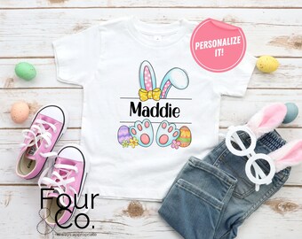 May include: White t-shirt featuring a blue bunny with pink ears and a yellow bow. The bunny is holding Easter eggs. The shirt says "Maddie" on it.  Personalise it!
