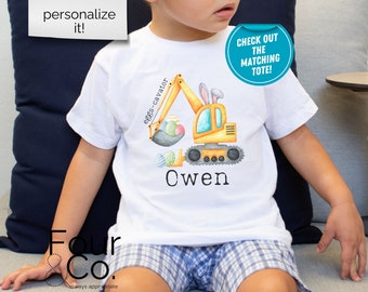 May include: A white t-shirt featuring a yellow excavator carrying Easter eggs. The excavator has bunny ears. The text "eggs-cavator" is written on the arm of the excavator. The name "Owen" is written below the excavator. The text "CHECK OUT THE MATCHING TOTE!" is in a blue circle on the right side of the image.