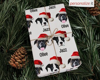 May include: White gift wrap featuring a repeating pattern of dogs wearing Santa hats, with the names "Olive" and "Jazz" printed on the wrapping paper. The gift is tied with twine.