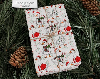May include: Wrapped gift with festive goose pattern. The wrapping paper features white geese wearing Santa hats and red jackets, holding candy canes and greenery. Tied with twine, the gift is set against a backdrop of pine needles and pine cones.