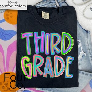 K&ouml;nnte beinhalten: Schwarzes T-Shirt mit den Worten "THIRD GRADE" in bunten, umrandeten Buchstaben. Das Shirt ist gefaltet und auf einer Jeans platziert. Die Worte "black comfort colors" stehen in einem wei&szlig;en Feld oben links.