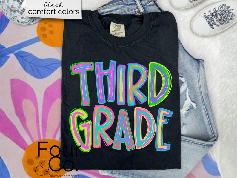 K&ouml;nnte beinhalten: Schwarzes T-Shirt mit den Worten "THIRD GRADE" in bunten, umrandeten Buchstaben. Das Shirt ist gefaltet und auf einer Jeans platziert. Die Worte "black comfort colors" stehen in einem wei&szlig;en Feld oben links.