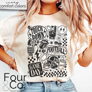 May include: Cream-colored t-shirt with a black and white graphic design featuring football-themed elements. The design includes the words "Touch Down", "Football Vibes", and "Game Day", along with footballs, a helmet, and a megaphone.