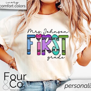 May include: A white t-shirt with the text "Mrs. Johnson FIRST grade" in rainbow colored crayon letters. The letters are outlined in black and have a small heart in the middle of the word "FIRST".