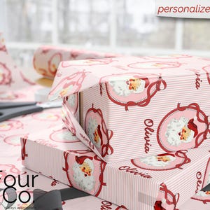 May include: Pink gift boxes wrapped in Santa Claus-themed paper with the name "Olivia" printed on them. The wrapping paper features a repeating pattern of Santa Claus in a decorative frame and vertical stripes. Scissors and a roll of wrapping paper are also visible.