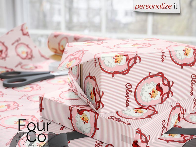 May include: Pink gift boxes wrapped in Santa Claus-themed paper with the name "Olivia" printed on them. The wrapping paper features a repeating pattern of Santa Claus in a decorative frame and vertical stripes. Scissors and a roll of wrapping paper are also visible.
