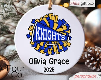May include: White ceramic ornament with a blue and yellow cheerleader design. The word "KNIGHTS" is displayed on a megaphone. The name "Olivia Grace" and the year "2025" are also printed on the ornament. A gold string is attached for hanging.