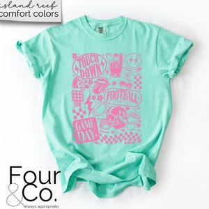 May include: A light turquoise t-shirt with a pink graphic design featuring football-related imagery and text. The design includes the words "TOUCH DOWN", "FOOTBALL", and "GAME DAY". The shirt is made of a soft material.