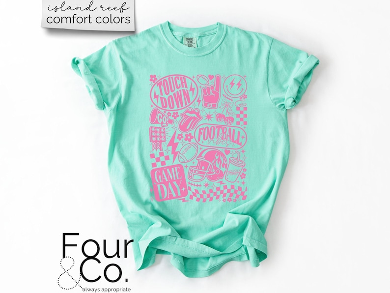 May include: A light turquoise t-shirt with a pink graphic design featuring football-related imagery and text. The design includes the words "TOUCH DOWN", "FOOTBALL", and "GAME DAY". The shirt is made of a soft material.