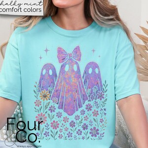 Puede incluir: Una camiseta azul claro con un dise&ntilde;o caprichoso de tres figuras de fantasmas y detalles florales. Los fantasmas son en tonos morados y rosas, con un lazo en el fantasma central. La camiseta tiene el texto "Four Co." en la parte inferior.