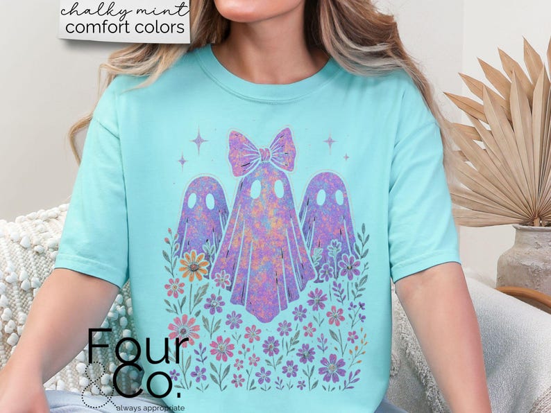 Puede incluir: Una camiseta azul claro con un dise&ntilde;o caprichoso de tres figuras de fantasmas y detalles florales. Los fantasmas son en tonos morados y rosas, con un lazo en el fantasma central. La camiseta tiene el texto "Four Co." en la parte inferior.
