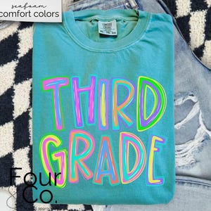 K&ouml;nnte beinhalten: Ein t&uuml;rkisfarbenes Comfort Colors T-Shirt mit den Worten "THIRD GRADE" in einer farbenfrohen, umrandeten Schriftart. Das Shirt ist gefaltet und auf einer hell gewaschenen Jeans platziert. Die Worte "seafoam comfort colors" befinden sich in der oberen linken Ecke.