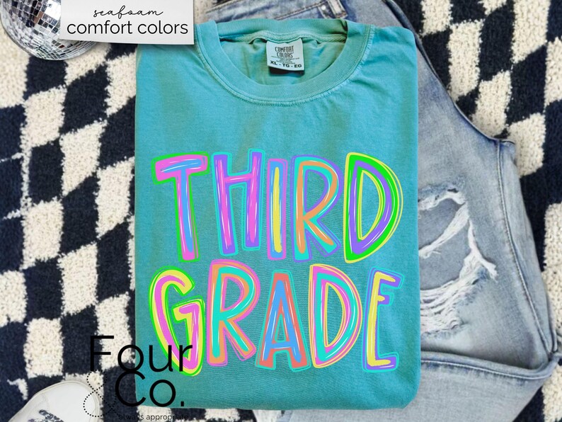K&ouml;nnte beinhalten: Ein t&uuml;rkisfarbenes Comfort Colors T-Shirt mit den Worten "THIRD GRADE" in einer farbenfrohen, umrandeten Schriftart. Das Shirt ist gefaltet und auf einer hell gewaschenen Jeans platziert. Die Worte "seafoam comfort colors" befinden sich in der oberen linken Ecke.