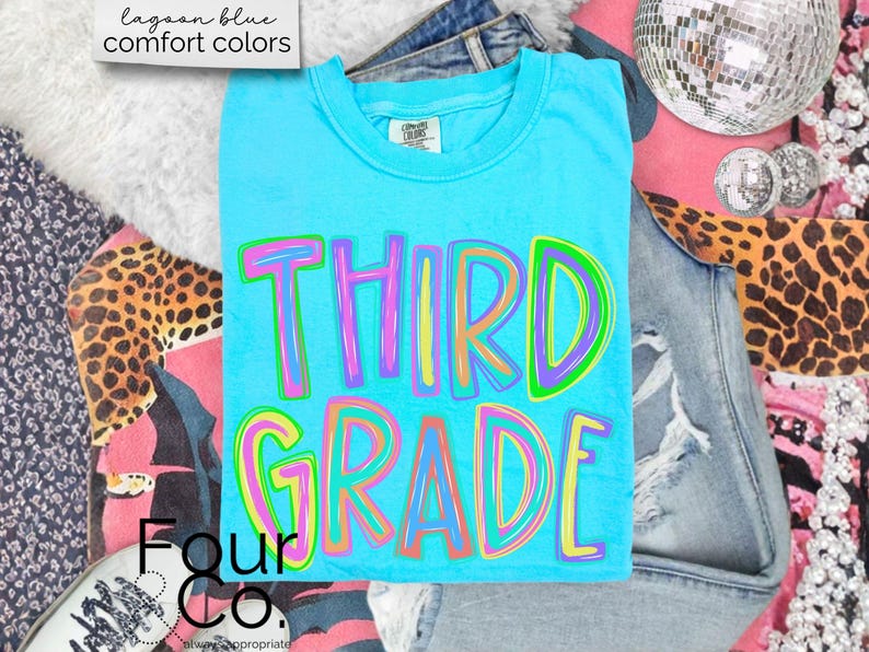 K&ouml;nnte beinhalten: Ein t&uuml;rkisblaues T-Shirt mit den Worten "THIRD GRADE" in bunten Buchstaben. Das Shirt ist von Jeans, einem Leopardenmuster und einer Discokugel umgeben.