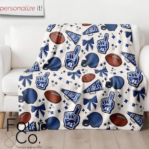 Könnte beinhalten: Eine cremefarbene Decke mit einem sich wiederholenden Muster aus Footballs, Helmen, Schaumstoffhänden, Schleifen und Sternen in Marineblau und Braun. Die Worte "GO EAGLES" sind ebenfalls enthalten. Die Decke ist über ein weißes Sofa drapiert.