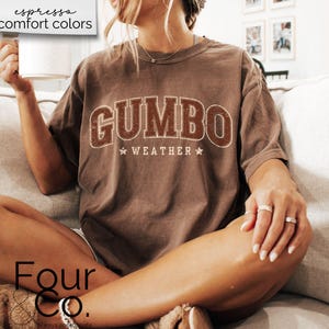 May include: A brown t-shirt with the text "GUMBO WEATHER" in a vintage collegiate style font. The shirt is worn by a person sitting on a couch.