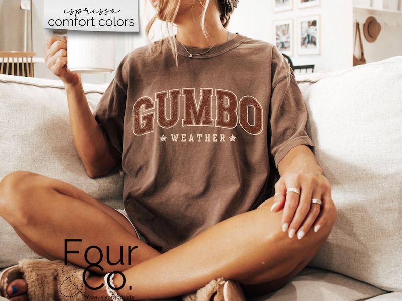 May include: A brown t-shirt with the text "GUMBO WEATHER" in a vintage collegiate style font. The shirt is worn by a person sitting on a couch.
