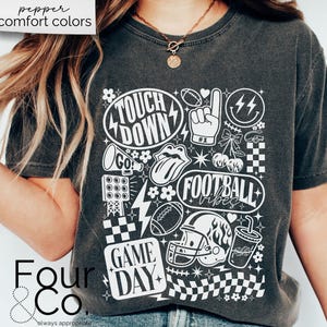 May include: Dark gray t-shirt with a white graphic design featuring football-themed elements like a helmet, football, and the words "Touchdown", "Football Vibes", and "Game Day". The shirt has the text "pepper comfort colors" at the top.