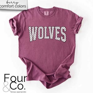 May include: A berry-colored t-shirt with the word "WOLVES" in white, polka-dotted letters. The shirt has rolled-up sleeves and the text "berry comfort colors" is in the upper left corner. The Four & Co. logo is in the lower left corner.