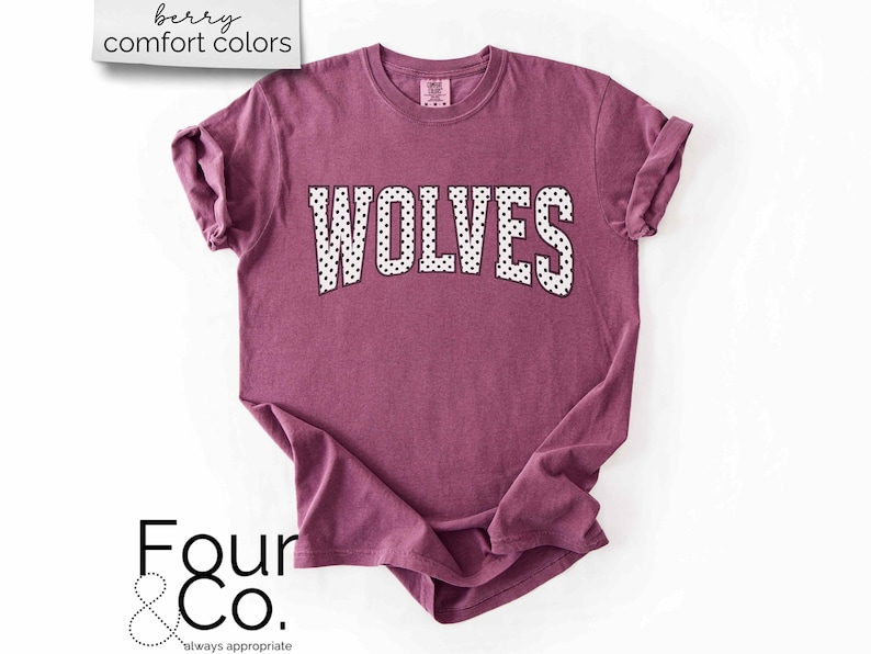 May include: A berry-colored t-shirt with the word "WOLVES" in white, polka-dotted letters. The shirt has rolled-up sleeves and the text "berry comfort colors" is in the upper left corner. The Four & Co. logo is in the lower left corner.