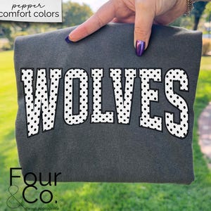 May include: Dark gray sweatshirt with the word "WOLVES" in large white letters with black polka dots. The sweatshirt is held up by a hand with purple nail polish. The words "pepper comfort colors" are in the top left corner.