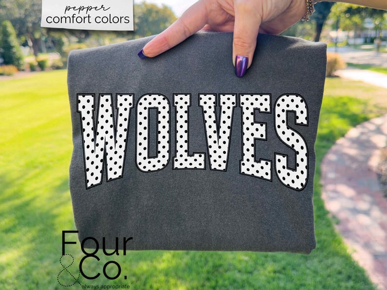 May include: Dark gray sweatshirt with the word "WOLVES" in large white letters with black polka dots. The sweatshirt is held up by a hand with purple nail polish. The words "pepper comfort colors" are in the top left corner.