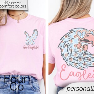 Pu&ograve; includere: T-shirt rosa chiaro con un design floreale di aquila. La parte anteriore presenta una piccola grafica di aquila e la scritta "Go Eagles!". Il retro mostra un'aquila pi&ugrave; grande con occhiali a forma di cuore e un papillon, con la parola "Eagles" sotto.
