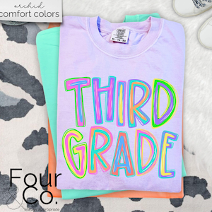 K&ouml;nnte beinhalten: Ein Stapel von drei T-Shirts in Pastellfarben: Lavendel, Mintgr&uuml;n und Pfirsich. Das oberste Shirt ist lavendelfarben und tr&auml;gt die bunten Buchstaben "THIRD GRADE". Die Worte "archid comfort colors" stehen oben links.