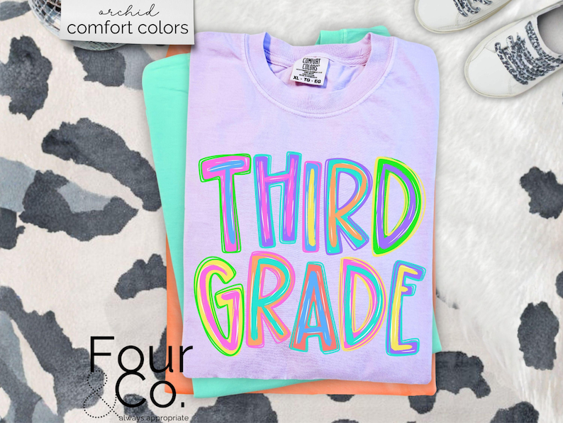 K&ouml;nnte beinhalten: Ein Stapel von drei T-Shirts in Pastellfarben: Lavendel, Mintgr&uuml;n und Pfirsich. Das oberste Shirt ist lavendelfarben und tr&auml;gt die bunten Buchstaben "THIRD GRADE". Die Worte "archid comfort colors" stehen oben links.