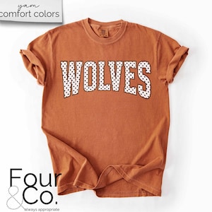 May include: A burnt orange t-shirt with the word "WOLVES" in white, dotted letters. The shirt has rolled-up sleeves and the text "yam comfort colors" is in the upper left corner. The logo "Four & Co." is in the lower left corner.