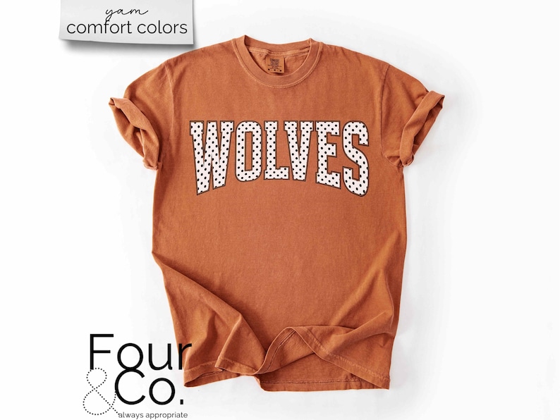 May include: A burnt orange t-shirt with the word "WOLVES" in white, dotted letters. The shirt has rolled-up sleeves and the text "yam comfort colors" is in the upper left corner. The logo "Four & Co." is in the lower left corner.