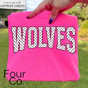 May include: A neon pink t-shirt with the word "WOLVES" in large, white, polka-dotted letters. The shirt is held up by a hand, and the text "neon pink comfort colors" is in the upper left corner. The brand logo "Four & Co." is at the bottom.