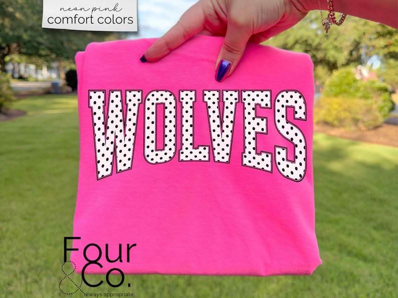 May include: A neon pink t-shirt with the word "WOLVES" in large, white, polka-dotted letters. The shirt is held up by a hand, and the text "neon pink comfort colors" is in the upper left corner. The brand logo "Four & Co." is at the bottom.