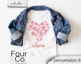 May include: White t-shirt with a red heart made of pink and red bows. The text "olivia" is written below the heart.