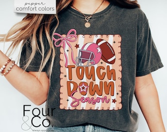 May include: Dark gray t-shirt with a football-themed graphic. The design features a pink football helmet and a brown football, with the words "Touch Down Season" in orange and pink. The shirt also has a pink bow.