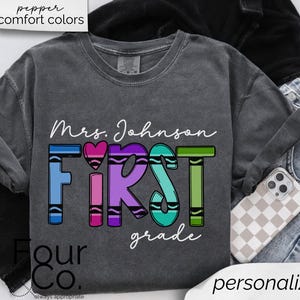 May include: A gray t-shirt with the text "Mrs. Johnson First Grade" in white lettering. The word "First" is made up of colorful crayon shapes.