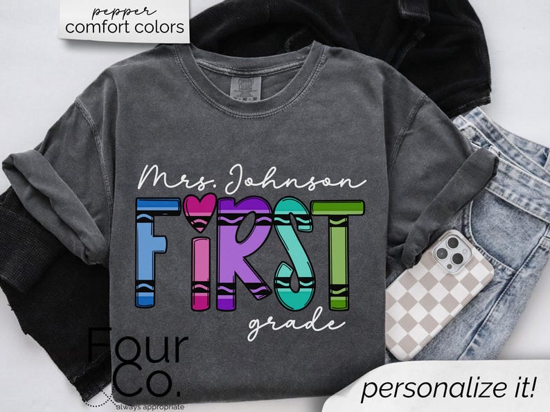 May include: A gray t-shirt with the text "Mrs. Johnson First Grade" in white lettering. The word "First" is made up of colorful crayon shapes.