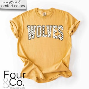 May include: Mustard-colored t-shirt with the word "WOLVES" in black and white polka dot letters. The shirt has rolled-up sleeves and a crew neck. The text "mustard comfort colors" is in the upper left corner, and the logo "Four & Co." is in the lower left corner.