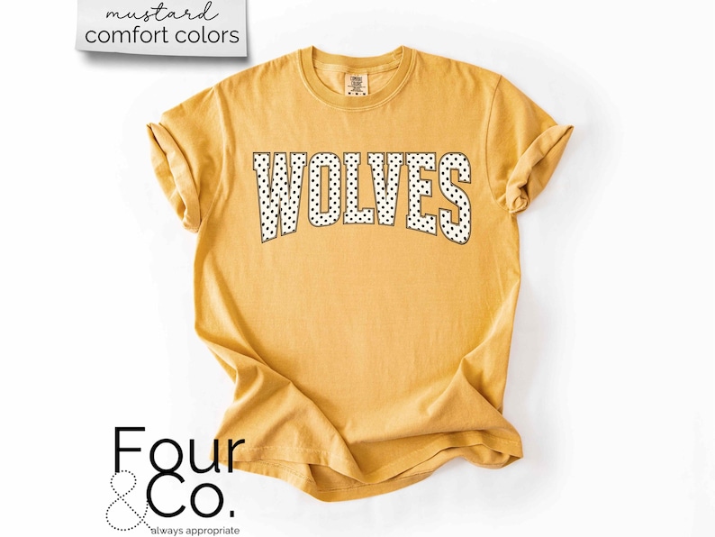 May include: Mustard-colored t-shirt with the word "WOLVES" in black and white polka dot letters. The shirt has rolled-up sleeves and a crew neck. The text "mustard comfort colors" is in the upper left corner, and the logo "Four & Co." is in the lower left corner.