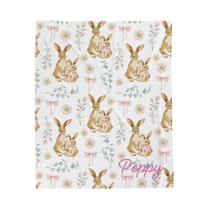 May include: A white baby blanket with a repeating pattern of brown bunnies holding pink baby bunnies, pink bows, and green leaves. The text "Poppy" is written in pink at the bottom of the blanket.