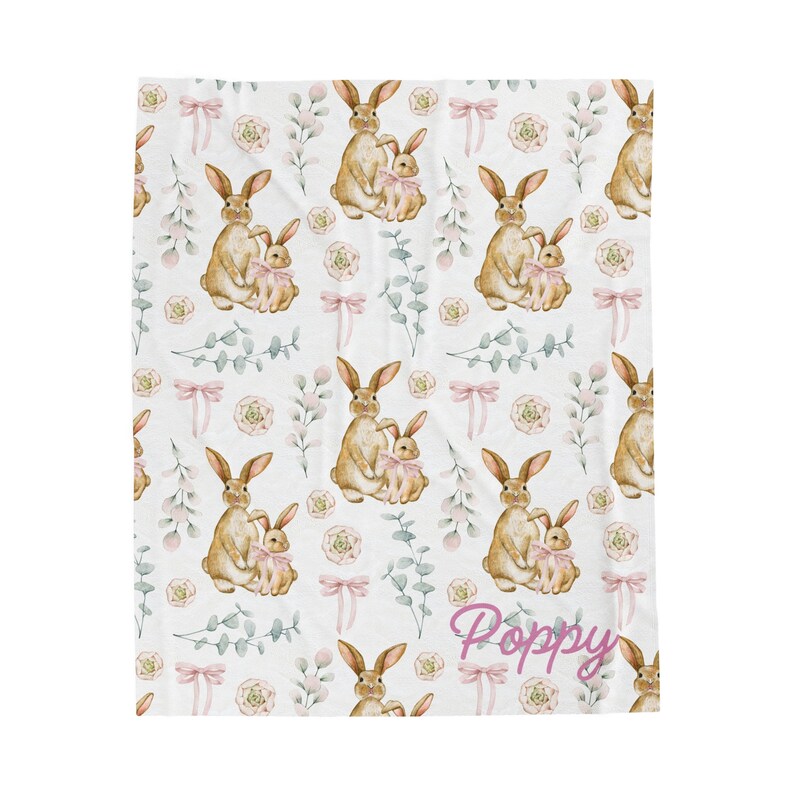 May include: A white baby blanket with a repeating pattern of brown bunnies holding pink baby bunnies, pink bows, and green leaves. The text "Poppy" is written in pink at the bottom of the blanket.